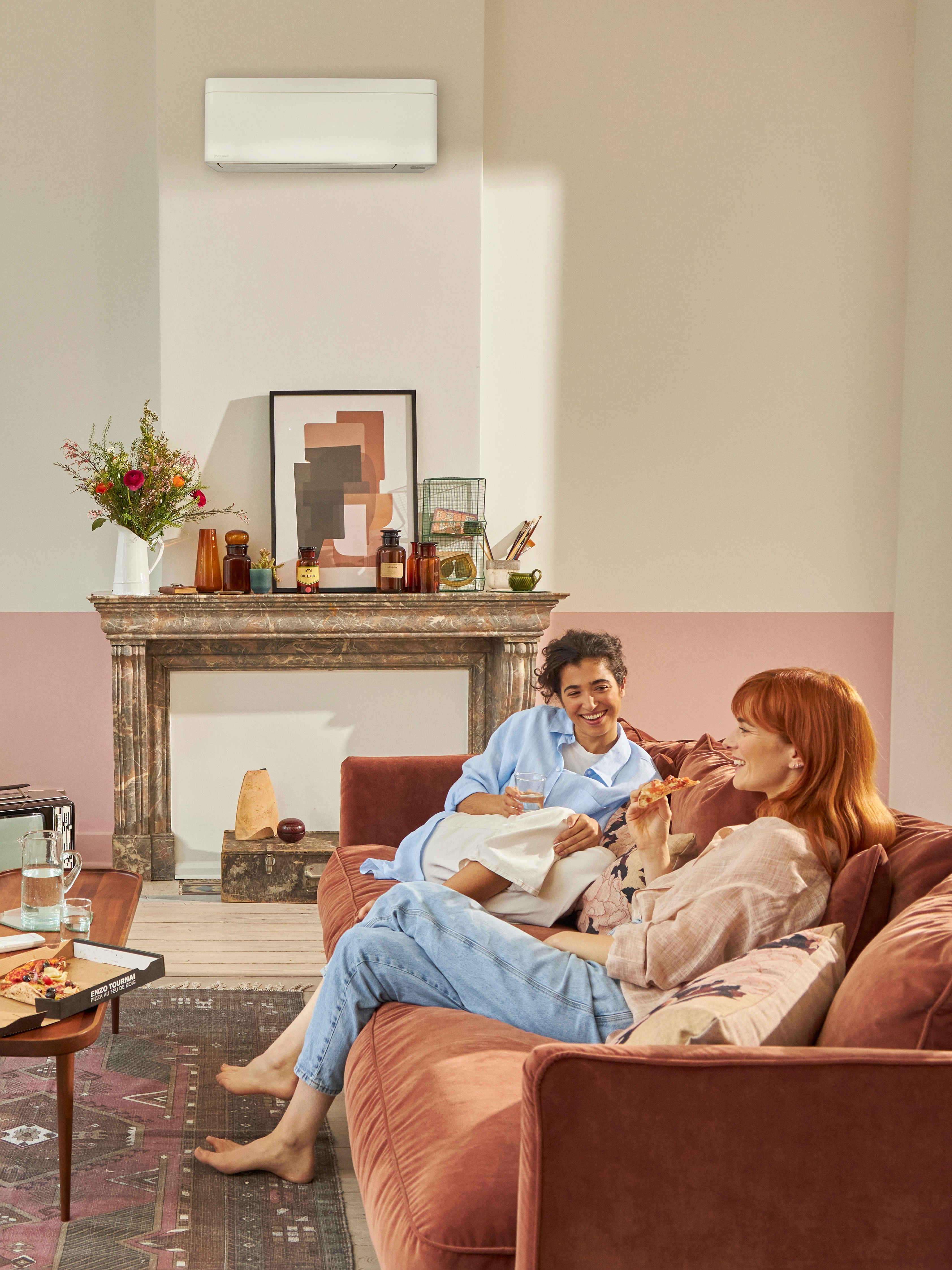 To kvinder sidder i en sofa i en smuk stue og taler sammen, mens de spiser takeaway-pizza.